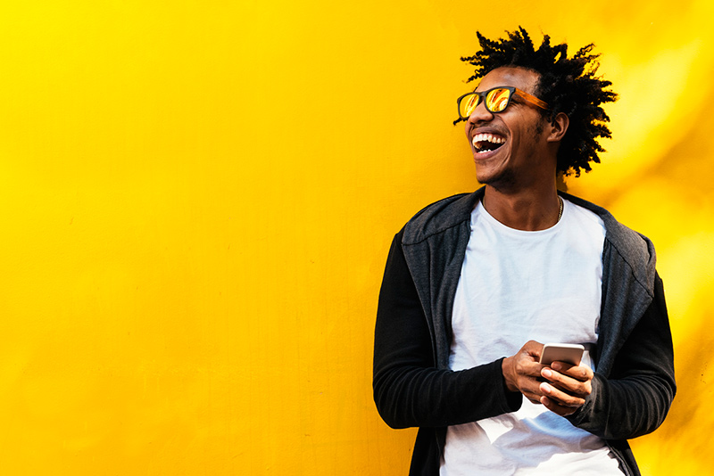 happy Black man wearing sunglasses using cell phone in front of bright yellow wall