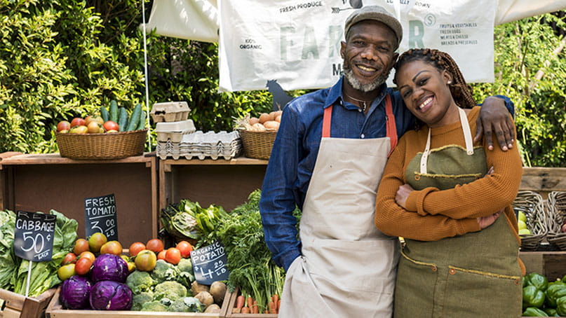 a Black man and woman wearing aprons are standing in front of their produce stand at a farmers market