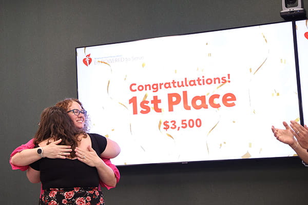 Katelyn France is hugging a facilitator at the ETS Minnesota Business Accelerator finale awards ceremony. A monitor in the background reads, "Congratulations! 1st Place, $3,500."