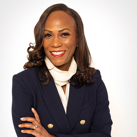 Pamela Garmon Johnson is standing with arms crossed and smiling on a white background. She is wearing a navy blazer with gold buttons and a white blouse.