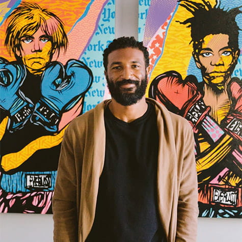 Ryan Mundy standing in front of colorful paintings of Andy Warhol and Jean-Michel Basquiat wearing boxing gloves