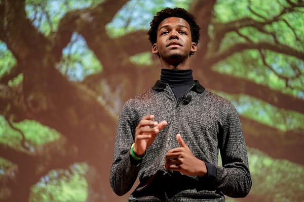 Russell Fearon, a Black man actively speaking with a large tree on a screen behind him