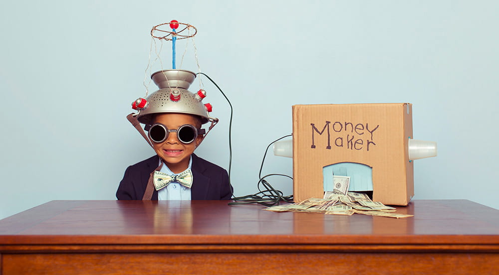 A young boy is sitting behind a desk in front of a light blue wall. He's wearing a suit jacket and bowtie with goggles and a metal pasta strainer on his head connected by wires to a cardboard box. The box has "Money Maker" written on it, and cash is piled under a cut-out in the box.