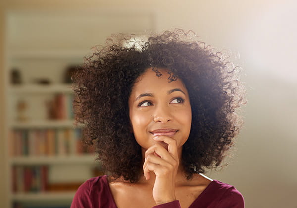 a Black woman in a thinking pose with her hand on her chin in front of a bookshelf