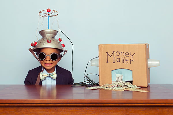 A young boy is sitting behind a desk in front of a light blue wall. He's wearing a suit jacket and bowtie with goggles and a metal pasta strainer on his head connected by wires to a cardboard box. The box has "Money Maker" written on it, and cash is piled under a cut-out in the box.