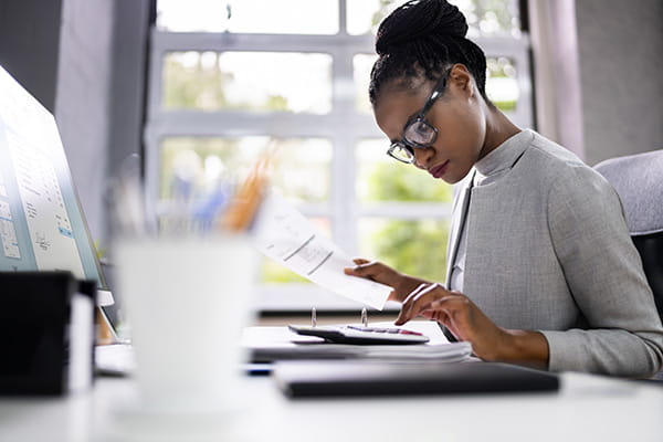 a Black woman in a grey turtleneck and glasses is sitting at a desk in front of a window using a calculator and holding an invoice a Black woman in a grey turtleneck and glasses is sitting at a desk in front of a window using a calculator and holding an invoice