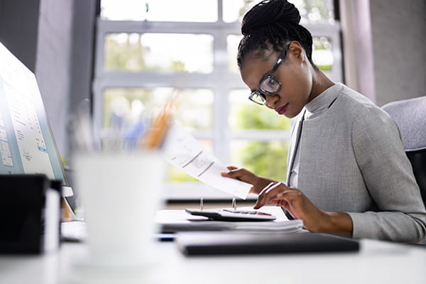 a Black woman in a grey turtleneck and glasses is sitting at a desk in front of a window using a calculator and holding an invoice