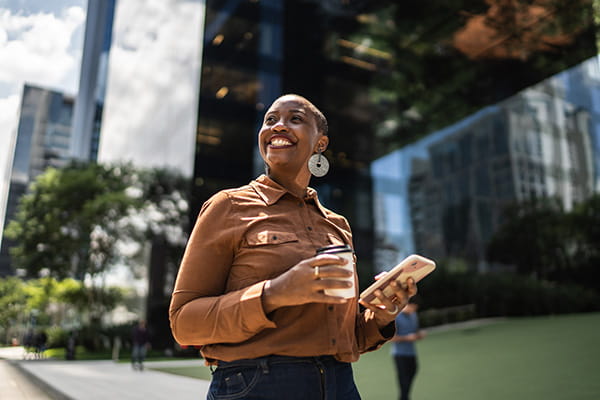 a Black woman is looking up smiling while holding a coffee and smartphone, walking on a city sidewalk with glass skyscrapers in the background