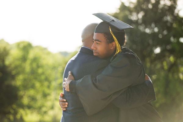 male graduate in cap and gown hugging person