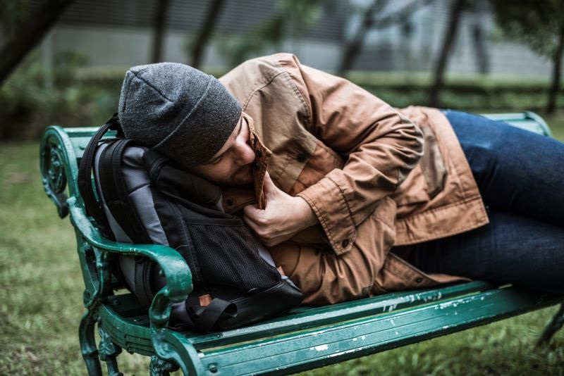 A man lying on a bench in a grassy area using a backpack as a pillow