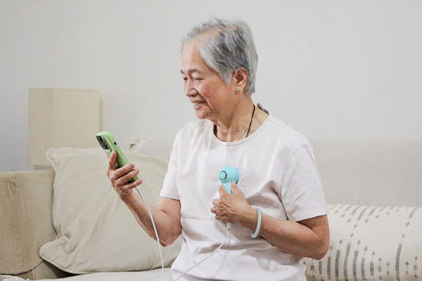 An older woman of Asian descent is using the SoundHeart electronic stethoscope with the Korion Health app on a smartphone from her sofa.